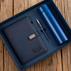 May include: A blue gift set in a blue box, including a notebook, pen, and a vacuum-insulated water bottle. The notebook has a silver clasp and a deer antler design with the initials "PM". The pen and water bottle are a matching blue color.