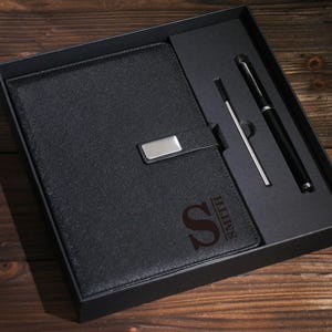 May include: A black gift set featuring a leather-bound notebook with a silver clasp, a pen, and a stylus. The notebook is embossed with the letter "S" and the name "SMITH". The set is presented in a black box.