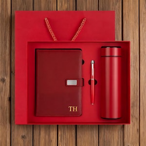 May include: A red gift set comprising a notebook with gold initials, a pen, and a flask. The set is presented in a red box and a matching gift bag, on a wooden surface. The notebook has a silver clasp.