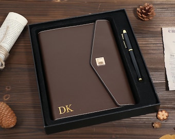 May include: A brown leather journal with a gold clasp and the initials "DK" in gold lettering, presented in a black gift box with a matching pen. The journal has white stitching.