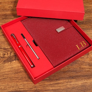 May include: A red gift set featuring a matching pen, refill, and a notebook with gold initials "LB". The set is presented in a red box, ideal for gifting. The notebook has a silver clasp.
