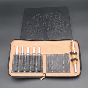 May include: A black leather pen case with a gold zipper, holding several silver-tipped pens and a wooden pen. A black embossed journal with a tree design is behind the case. The case has a tan interior.
