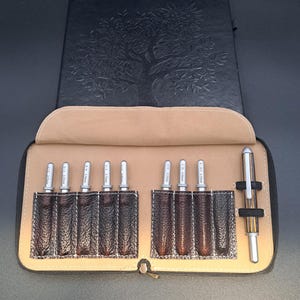 May include: A black leather-like journal with an embossed tree design sits behind an open tan and dark brown textured case. The case contains eight silver metal crochet hooks with size markings and one striped pen held by an elastic band.