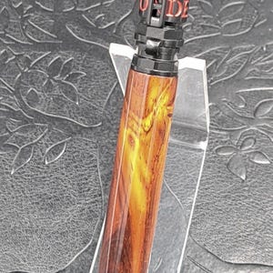 May include: A ballpoint pen featuring a polished wooden barrel with natural grain patterns, paired with a black metal cap and tip. The cap displays a firefighter helmet emblem and red text that reads "HONOR", "REVERED", "DUTY", and "DEEDS".