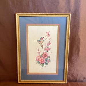 以下が含まれることがあります： ピンクの花の近くにいるハチドリを描いた額入りのアートワーク。鳥は緑、赤、白の羽を持っています。アートワークは、クリーム色の背景に青とピンクのマットがあり、金色のフレームに収められています。