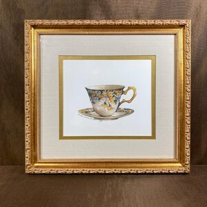 Puede incluir: Una pintura en acuarela enmarcada de una taza de té y un platillo con un patrón floral en tonos amarillos y azules. La obra está dentro de un marco dorado decorativo con un paspartú color crema y un borde interior dorado fino.