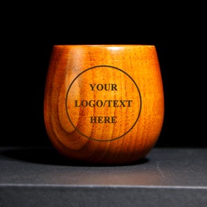 Puede incluir: Una copa de vino sin tallo de madera con un acabado de veta de madera natural. La copa tiene forma redonda y presenta el texto "YOUR LOGO/TEXT HERE" dentro de un círculo.
