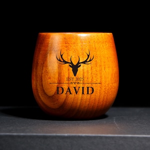 以下が含まれることがあります： 天然仕上げの木製タンブラー。タンブラーには、テキスト「DAVID」と「EST. 2025」の上に黒い鹿の頭のグラフィックが描かれています。カップは丸みを帯びたベースと広い開口部で設計されています。