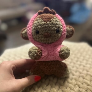 May include: A handmade crochet monkey doll with a pink hooded jacket. The monkey has brown and tan fur, black eyes, and small ears. The doll is sitting on a textured surface.
