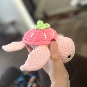 May include: A handmade crochet turtle toy in shades of pink and green. The turtle's shell is a vibrant pink, resembling a strawberry, with white stitch details. The head, flippers, and body are a soft pink. The toy is designed with black safety eyes.