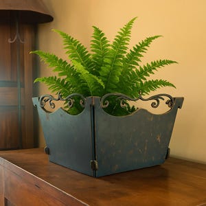 Puede incluir: Una jardinera cuadrada de metal azul oscuro con un acabado envejecido, que contiene un helecho verde vibrante. La jardinera tiene adornos decorativos a lo largo del borde superior y está colocada sobre una superficie de madera. La estética general es rústica y elegante.
