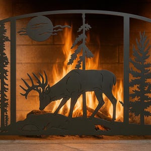 Puede incluir: Pantalla de chimenea de metal negro con una silueta de ciervo, árboles y un diseño de sol. La pantalla tiene una parte superior arqueada y está colocada sobre un fondo de chimenea encendida.