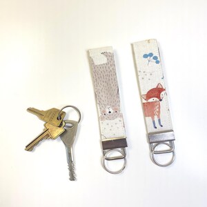 Peut inclure: Deux porte-clés en tissu avec des ferrures en métal argenté et des anneaux fendus. L'un présente un motif d'ours clair, l'autre un motif de renard avec des baies bleues. Un jeu de trois clés en métal est attaché à l'un des anneaux fendus.