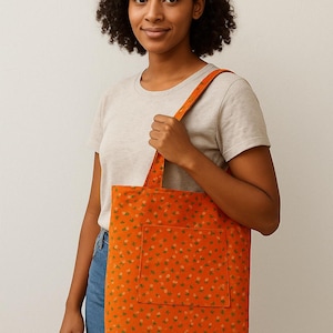 May include: Orange tote bag with a floral pattern. The bag has a large front pocket and a matching strap. The floral pattern features small green and white flowers on an orange background. The tote bag is made of fabric.