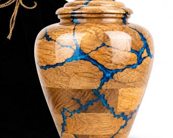 May include: A cremation urn crafted from segmented light brown wood with a polished finish, featuring vibrant blue resin veins running through the structure. The urn has a tiered lid and is accompanied by a black velvet drawstring storage bag.