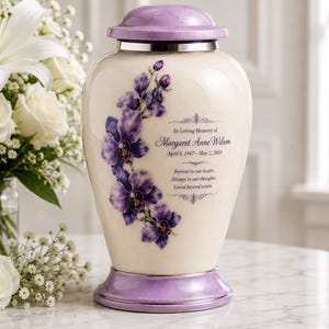 May include: A cream-colored urn with a lavender lid and base, adorned with purple orchid floral designs. The urn has text that reads "In Loving Memory of Margaret Anne Wilson, April 5, 1947 - May 2, 2024." A bouquet of white flowers is in the background.