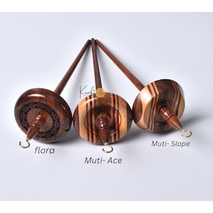 Op de afbeelding: Drie houten drop spindles met verschillende ontwerpen. De spindles hebben een donkerbruine schacht en een ronde, decoratieve basis. De basis van elke spindle is gelabeld met een naam: "flora", "Muti-Ace" en "Muti-Slope". Elke spindle heeft een metalen haak.