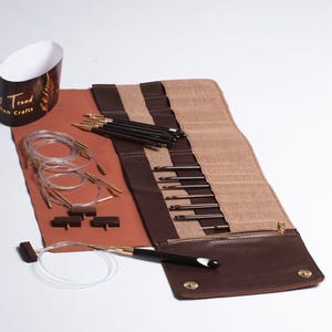 Puede incluir: Un estuche de agujas de tejer de cuero marrón y tela beige con varias agujas, cables y accesorios. El estuche está abierto, mostrando las agujas y otras herramientas. También se ve un recipiente de papel con el texto "Trendy Crafts".