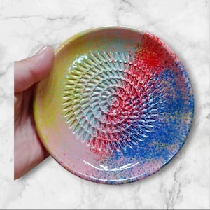 Może przedstawiać: Kolorowy ceramiczny talerz do tarcia z spiralną powierzchnią do tarcia. Talerz ma żywą mieszankę kolorów, w tym żółty, różowy, czerwony i niebieski, z efektem nakrapianej farby. Talerz jest trzymany przez dłoń.