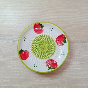Może przedstawiać: Biała ceramiczna tarka z zielonym środkiem i obramowaniem, ozdobiona ilustracjami czerwonych pomidorów. Tarka ma teksturowaną powierzchnię do ścierania i jest przeznaczona do użytku w kuchni.