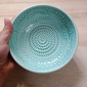 Może przedstawiać: Turkusowa ceramiczna miska do tarcia z teksturowanym środkiem i dekoracyjnymi zawijasami na obrzeżu. Miskę trzyma ręka i stoi na jasnej drewnianej powierzchni. Misa jest przeznaczona do tarcia.