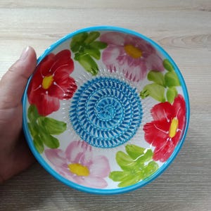 Może przedstawiać: Ceramiczna miska z kwiatowym wzorem. Misa ma czerwone i różowe kwiaty z żółtymi środkami, zielone liście i niebieską obwódkę. Środek miski ma teksturowany niebieski wzór.