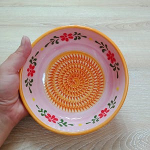 Może przedstawiać: Różowa ceramiczna tarka z pomarańczowym środkiem i kwiatowym wzorem. Tarka ma żółtą obwódkę i jest ozdobiona czerwonymi kwiatami i zielonymi liśćmi. Tarka jest trzymana w dłoni.