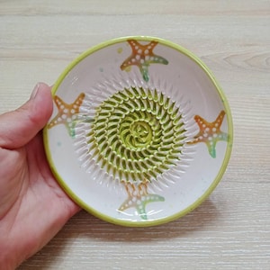 Może przedstawiać: Biała ceramiczna tarka z zieloną i żółtą spiralną powierzchnią do ścierania. Obręcz jest żółta, a miska ozdobiona pomarańczowymi i zielonymi rozgwiazdami. Tarka jest trzymana w dłoni.