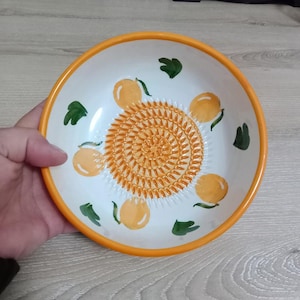 Może przedstawiać: Biała ceramiczna miska do tarcia z pomarańczową obwódką i spiralną tarką pośrodku. Miska jest ozdobiona pomarańczowymi owocami i zielonymi liśćmi. Miskę trzyma ręka.