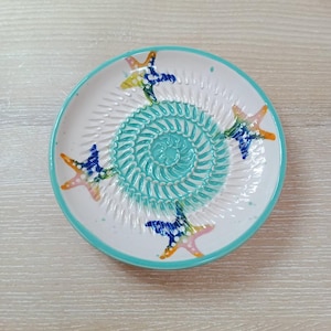 Może przedstawiać: Biały ceramiczny talerz do tarcia z turkusowym spiralnym środkiem i turkusową obwódką. Talerz posiada kolorowe wzory rozgwiazd w kolorze żółtym, pomarańczowym, różowym i niebieskim. Talerz jest przeznaczony do tarcia żywności.