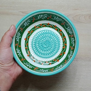 Może przedstawiać: Ceramiczna miska do tarcia z turkusowym wnętrzem i dekoracyjnym zielono-białym obramowaniem. Misa posiada spiralną powierzchnię do tarcia pośrodku i jest ozdobiona pomarańczowymi detalami. Misa jest trzymana w dłoni.