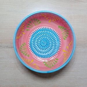 Może przedstawiać: Ręcznie malowana ceramiczna miska do tarcia. Miska ma różowe wnętrze z niebieską spiralną tarką pośrodku. Obręcz jest niebieska i ozdobiona żółtymi i zielonymi kwiatowymi akcentami. Dekoracyjny artykuł kuchenny.