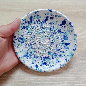 Może przedstawiać: Biała ceramiczna tarka do czosnku o teksturowanej powierzchni i spiralnym wzorze. Tarka jest ozdobiona niebieskimi i turkusowymi plamami farby. Okrągła tarka jest trzymana w dłoni.