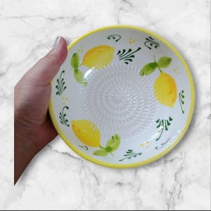 Może przedstawiać: Biała ceramiczna miska do tarcia z żółtą obwódką i cytrynowym wzorem. Misa posiada centralną powierzchnię do tarcia i jest ozdobiona żółtymi cytrynami i zielonymi liśćmi. Dekoracyjny artykuł kuchenny.