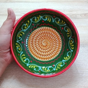Może przedstawiać: Ceramiczna miska do tarcia o żywym wzorze. Miska ma zielone wnętrze ze spiralnym wzorem pośrodku i dekoracyjnymi wirami. Obręcz jest czerwona, a miska jest trzymana na jasnej drewnianej powierzchni.