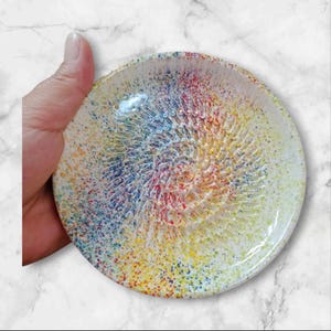 Może przedstawiać: Okrągła tarki ceramiczna z teksturowaną powierzchnią i kolorowym, nakrapianym wzorem. Tarka ma spiralny wzór pośrodku, z gradientem kolorów, w tym żółtym, niebieskim i czerwonym. Obręcz jest biała.