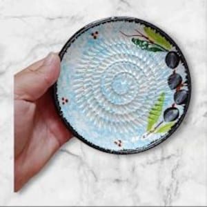 Może przedstawiać: Mały, okrągły ceramiczny talerz do tarcia z jasnoniebieskim środkiem i czarną obwódką. Talerz posiada spiralną powierzchnię do tarcia, malowane oliwki i zielone liście. Talerz jest trzymany przez dłoń na tle marmuru.