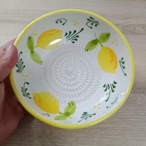 Może przedstawiać: Biała ceramiczna miska do tarcia z żółtą obwódką i cytrynowym wzorem. Miska posiada teksturowaną powierzchnię do tarcia pośrodku i jest ozdobiona żółtymi cytrynami i zielonymi liśćmi. Miska jest przeznaczona do użytku w kuchni.