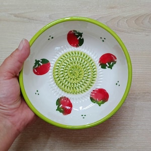 Może przedstawiać: Biała ceramiczna miska do tarcia z zieloną obwódką i zieloną powierzchnią do tarcia pośrodku. Miska jest ozdobiona ilustracjami czerwonych pomidorów i małymi zielonymi kropkami. Miska jest trzymana przez dłoń.
