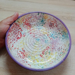Może przedstawiać: Ceramiczna miska do tarcia z białym wnętrzem i fioletową obwódką. Wnętrze miski zdobią kolorowe plamy czerwonej, niebieskiej, żółtej i zielonej farby. Środek miski ma teksturowaną powierzchnię do tarcia.