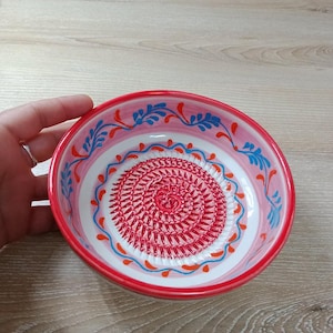 Może przedstawiać: Mała, okrągła ceramiczna miska z czerwonym rantem i dekoracyjnym wnętrzem. Misa ma spiralny wzór pośrodku, otoczony niebieskimi i pomarańczowymi kwiatowymi wzorami na białym i różowym tle. Misa jest trzymana przez dłoń.