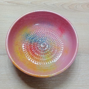 Peut inclure: Un bol râpe en céramique rose avec un intérieur aux couleurs de l'arc-en-ciel. Le centre du bol présente une surface de râpage texturée, entourée d'un dégradé de jaune, d'orange, de rouge, de bleu et de violet. Le bol est conçu pour la cuisine.
