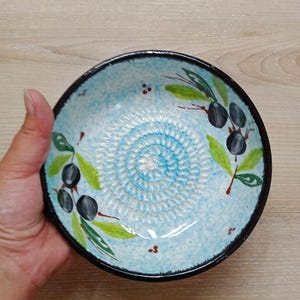 Może przedstawiać: Ręcznie malowana ceramiczna miska do tarcia oliwek z jasnoniebieskim wnętrzem i czarną obwódką. Miska posiada spiralną powierzchnię do tarcia i jest ozdobiona motywami gałązek oliwnych w kolorze zielonym i czarnym.