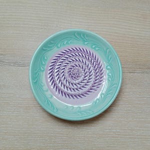 Może przedstawiać: Ceramiczny talerz do tarcia z jasnoturkusową obwódką i lawendowym środkiem. Środek ma spiralny wzór wypukłych grzbietów do tarcia. Obrzeże ma dekoracyjny wytłoczony wzór.