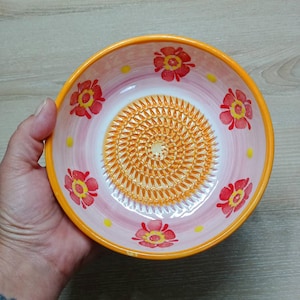 Może przedstawiać: Ceramiczna miska do tarcia z kwiatowym wzorem. Miska ma różowo-białe wnętrze z pomarańczową powierzchnią do tarcia. Czerwone kwiaty i żółte kropki zdobią obręcz. Obręcz jest pomarańczowa.