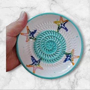 Może przedstawiać: Biała ceramiczna tarka z turkusowym spiralnym wzorem pośrodku i turkusową obwódką. Tarka posiada kolorowe wzory rozgwiazd na obrzeżu. Ręka trzyma tarkę.