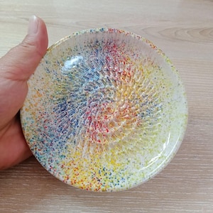 Może przedstawiać: Okrągła tarki ceramiczna z kolorowym, nakrapianym wzorem. Środek ma teksturowany, spiralny wzór z mieszanką niebieskiego, czerwonego, żółtego i białego. Obręcz jest błyszcząco biała. Średnica ok. 12 cm.