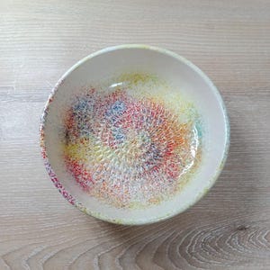 Może przedstawiać: Ręcznie robiona ceramiczna miska z teksturowanym wnętrzem. Misa ma nakrapiany wzór z żywą mieszanką kolorów, w tym czerwonym, żółtym, niebieskim i zielonym. Obręcz jest w kolorze miękkiej śmietanki, a miska stoi na jasnej drewnianej powierzchni.