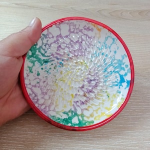 Może przedstawiać: Ceramiczna tarkownica do czosnku z czerwoną obwódką. Wnętrze posiada spiralną powierzchnię do ścierania i kolorowy wzór akwarelowy w odcieniach zieleni, niebieskiego, fioletu i żółci. Tarkownica jest trzymana w dłoni.