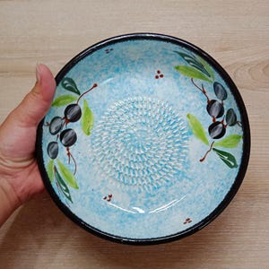 Może przedstawiać: Ręcznie malowana ceramiczna miska do tarcia oliwy z oliwek. Miska ma jasnoniebieskie wnętrze z teksturowaną powierzchnią do tarcia pośrodku. Czarne oliwki i zielone liście są namalowane wokół krawędzi. Obręcz jest czarna.